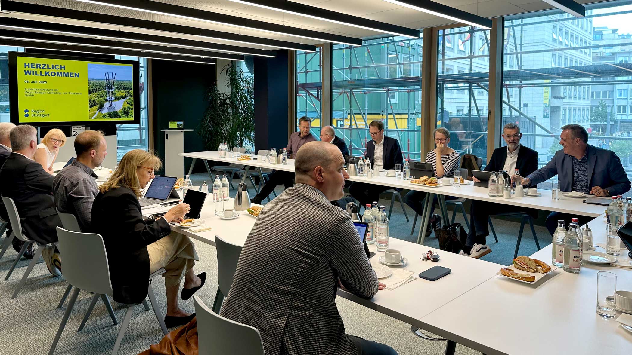 Personen in Business-Kleidung sitzen an Tischen, die U-förmig in Richtung eines großen Bildschirms ausgerichtet sind.