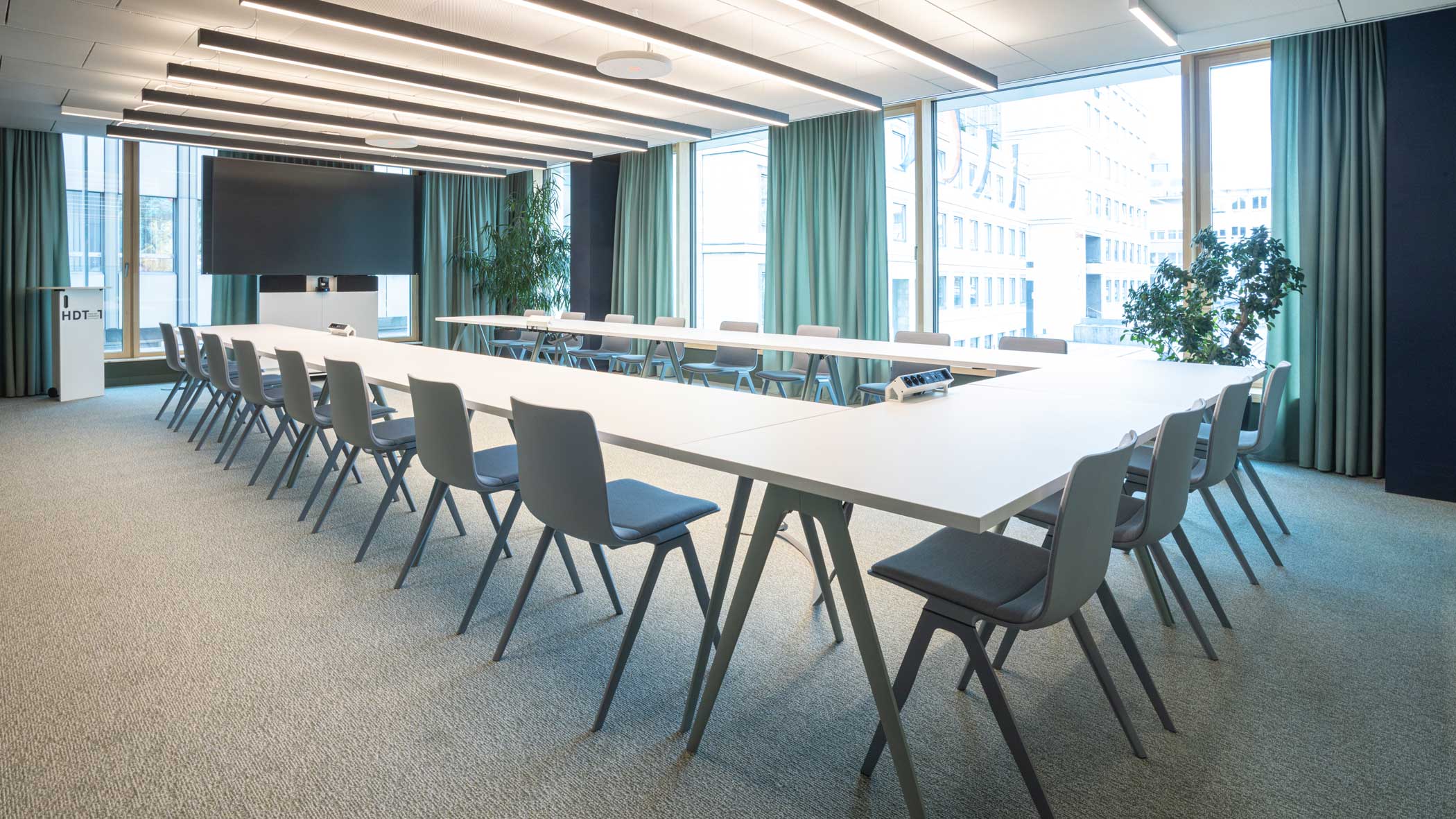 Klassischer Konferenzraum in gedeckten Farben mit U-förmig gestellten weißen Tischen und Bestuhlung. An der Stirnseite befindet sich ein großes Bildschirm.
