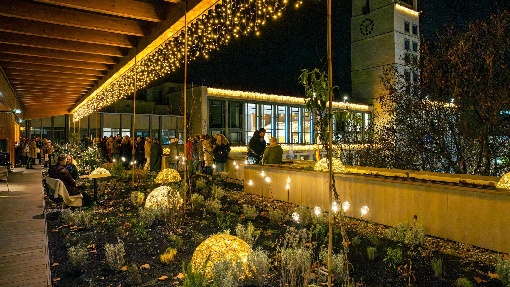 Dachterrasse des Haus des Tourismus mit vielen Lichtern: beleuchtete geflochtene Halbkugeln im Blumenbeet, Lichterkette am Dach und Leuchtkugeln auf Stäben. Im Hintergrund stehen Personen in warmer Kleidung zusammen.