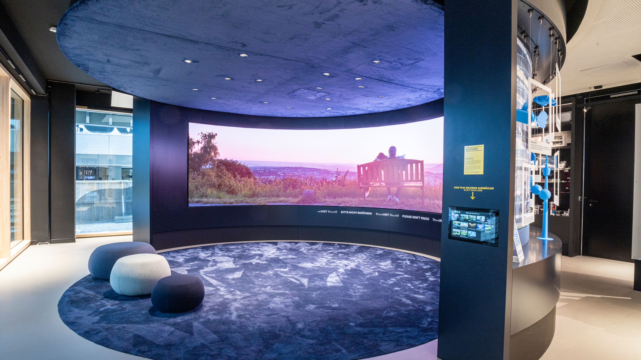 Wand im Halbkreis mit Bildschirm. Davor ein runder Teppich, auf dem drei verschieden große Poufs als Sitzgelegenheiten dienen.