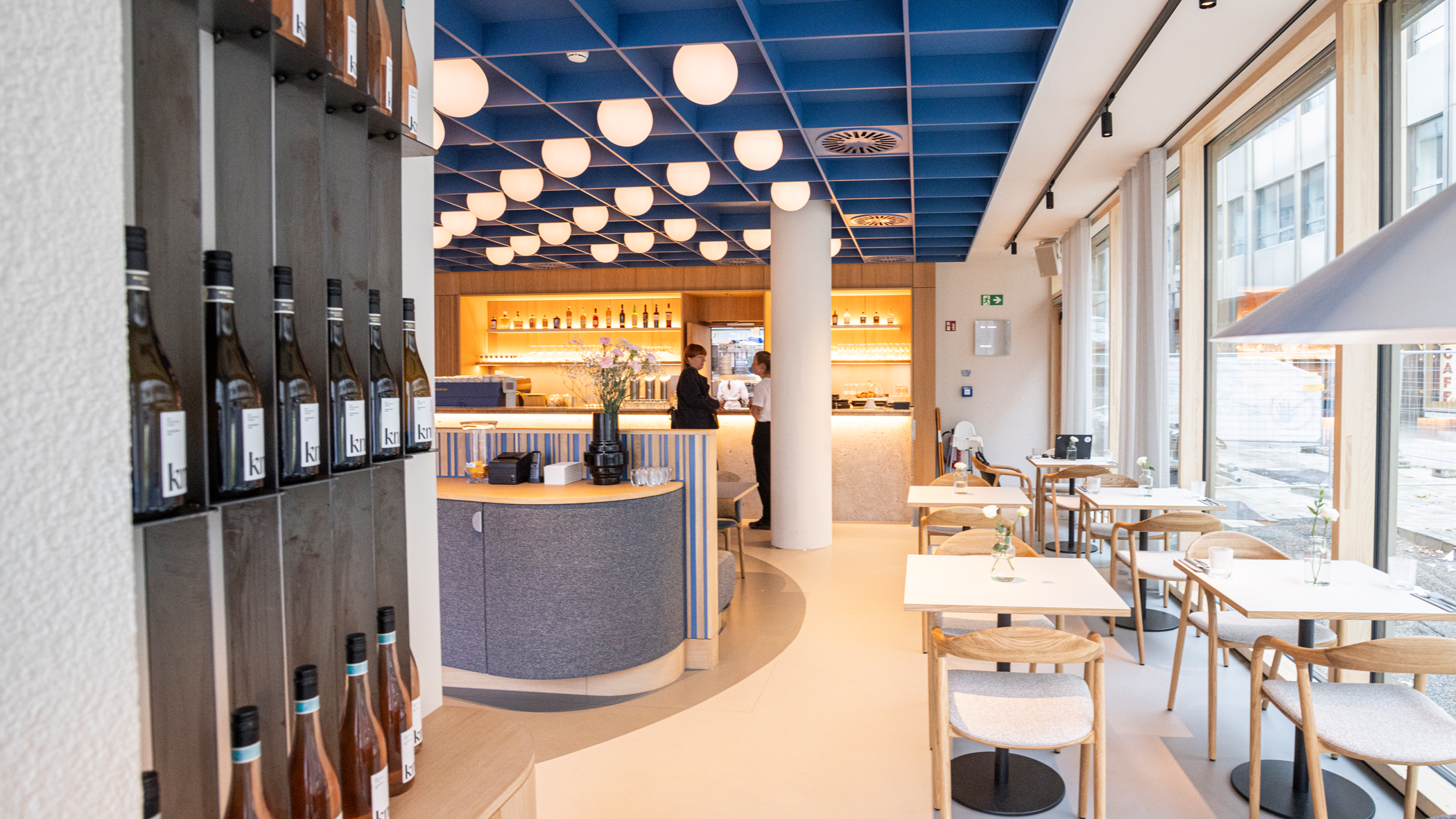 Weinregal auf der linken Seite, geradeaus die Theke, rechts an den bodentiefen Fenstern kleine Tische mit je zwei Stühlen.