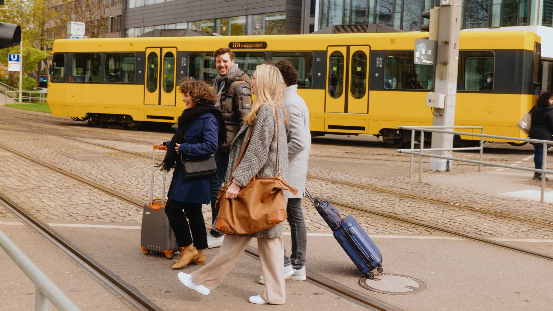 Eine Gruppe junger Leute mit Gepäck überquert eine Straße, im Hintergrund durchquert eine gelbe SSB U-Bahn das Bild.