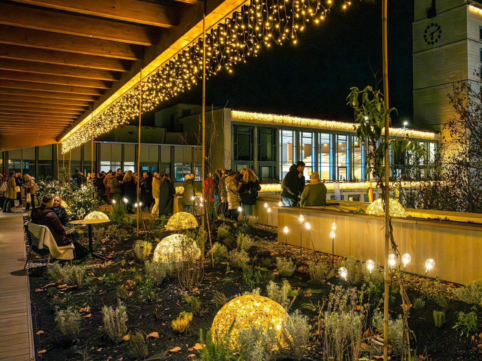 Dachterrasse zur Weihnachtszeit, SMG, Sarah Schmid Dachterrasse des Haus des Tourismus mit vielen Lichtern: beleuchtete geflochtene Halbkugeln im Blumenbeet, Lichterkette am Dach und Leuchtkugeln auf Stäben. Im Hintergrund stehen Personen in warmer Kleidung zusammen.