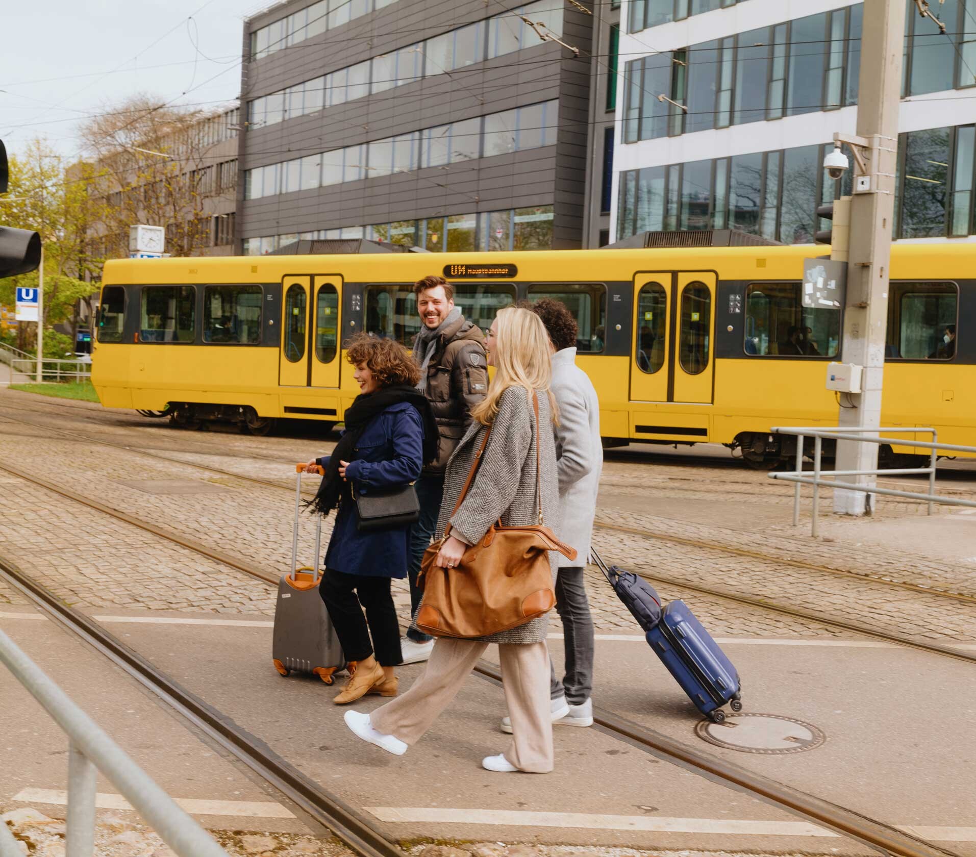 Eine Gruppe junger Leute mit Gepäck überquert eine Straße, im Hintergrund durchquert eine gelbe SSB U-Bahn das Bild.