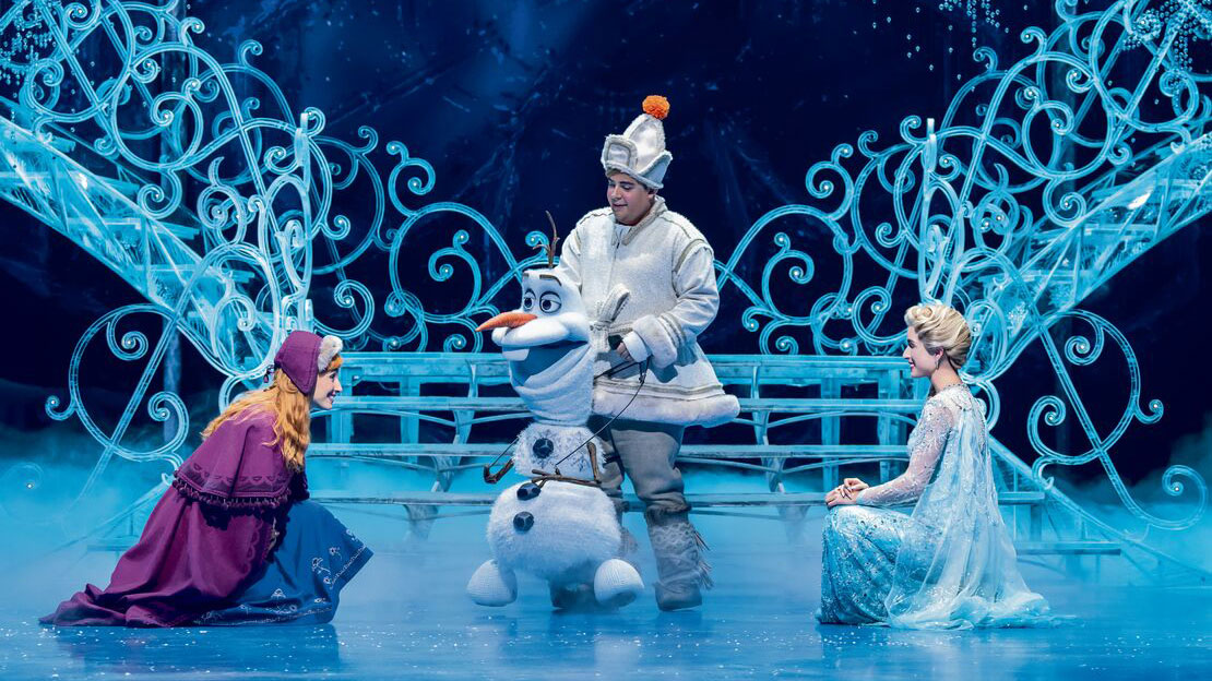 Die Schwestern Elsa und Anna knien auf dem Boden. In ihrer Mitte der Schneemann Olaf. Im Hintergrund eine geschwungene gefrorene Treppe.